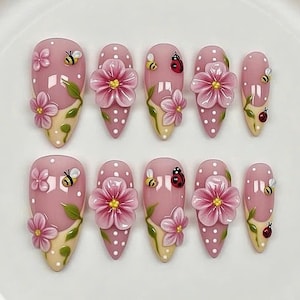 May include: Set of ten artificial nails in a light pink color with white polka dots. Each nail features a floral design with a 3D flower, a bee, and a ladybug. The base of each nail is a light yellow color.