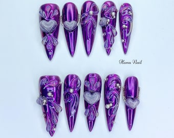 Puede incluir: Un conjunto de diez uñas artificiales largas y puntiagudas de color púrpura. Cada uña presenta un diseño de corazón plateado, adornado con pequeñas cuentas plateadas y detalles decorativos. Las uñas tienen un acabado brillante y están dispuestas sobre una superficie blanca. El texto "Hana Nail" es visible.