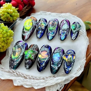 May include: A set of ten artificial nails with a unique design. Each nail features a dark blue base with iridescent accents and a clear, textured overlay. The nails are arranged on a white, lace-edged surface.