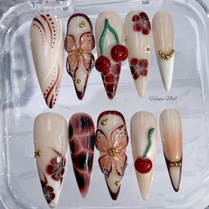 May include: A set of ten almond-shaped artificial nails with various designs. The nails feature floral and cherry designs in red, white, and gold, with the text "Hana Nail" visible on one nail. The nails are displayed in a clear plastic case.