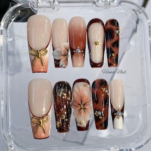 May include: A set of ten artificial nails in a clear plastic case. The nails feature various designs, including a bow, floral accents, and a leopard print. The color palette includes nude, brown, and gold tones. The text "Hana Nail" is visible.