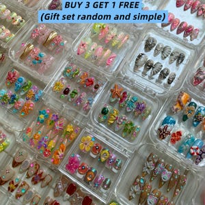May include: A collection of artificial nails in clear plastic cases. The nails feature various designs, including floral patterns, glitter, and 3D embellishments. The text "BUY 3 GET 1 FREE" is visible at the top.