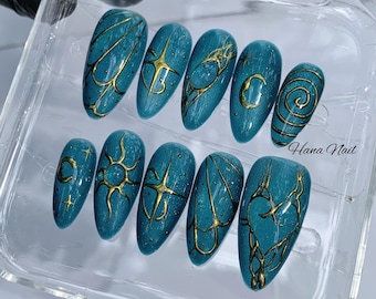May include: A collection of ten teal-coloured press-on nails with gold abstract designs. Each nail has a unique pattern, including lines, stars, and swirls. The nails are presented in a clear plastic container. The text "Hana Nail" is visible.