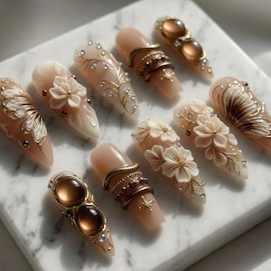 May include: A collection of ten almond-shaped artificial nails. Each nail features intricate designs, including floral accents, gold filigree, and embedded gemstones. The nails are primarily nude with gold, brown, and white details, creating an elegant aesthetic.