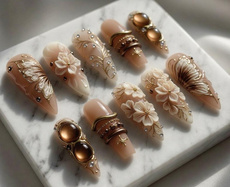 May include: A collection of ten almond-shaped artificial nails. Each nail features intricate designs, including floral accents, gold filigree, and embedded gemstones. The nails are primarily nude with gold, brown, and white details, creating an elegant aesthetic.