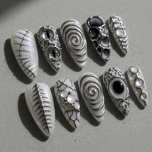 May include: A collection of ten artificial nails with intricate designs. The nails feature silver embellishments, including swirls, eye-like shapes, and geometric patterns, set against a white base. Some nails have a textured, metallic appearance, while others incorporate pearl-like accents.
