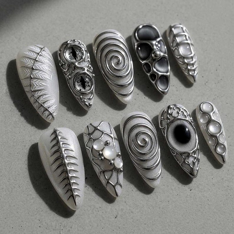 May include: A collection of ten artificial nails with intricate designs. The nails feature silver embellishments, including swirls, eye-like shapes, and geometric patterns, set against a white base. Some nails have a textured, metallic appearance, while others incorporate pearl-like accents.