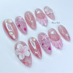 Peut inclure: Ensemble de dix faux ongles roses et blancs avec diff&eacute;rents motifs sur le th&egrave;me de l'&eacute;t&eacute;, notamment des coquillages, des &eacute;toiles de mer et des fleurs. Les ongles sont d&eacute;cor&eacute;s de paillettes et de strass. "Hana Nail" est &eacute;crit dans le coin sup&eacute;rieur droit de l'image.