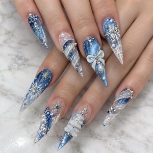 May include: Long stiletto-shaped nails featuring a blue and white color palette. The design includes intricate white snowflake patterns, raised 3D white bows, small pearls, silver rhinestones, and a textured 3D white Christmas tree accent.