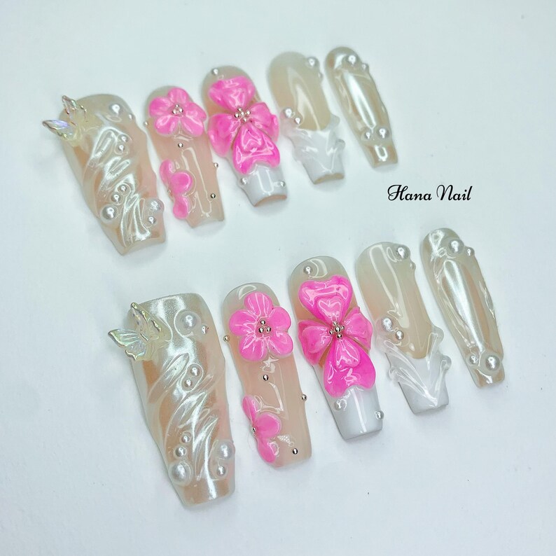 Puede incluir: Un conjunto de diez u&ntilde;as artificiales con diversos dise&ntilde;os. Algunas u&ntilde;as presentan un acabado nacarado con detalles de perlas y adornos de mariposas. Otras tienen dise&ntilde;os de flores y lazos rosas brillantes, y puntas blancas. El texto "Hana Nail" es visible.