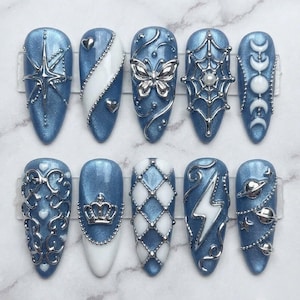 May include: A set of ten almond-shaped press-on nails featuring a metallic blue base with white accents. The designs include silver 3D charms such as a star, hearts, a butterfly, a spider web with a pearl, moon phases, filigree, a crown, a lattice pattern, a lightning bolt, and planets with stars.