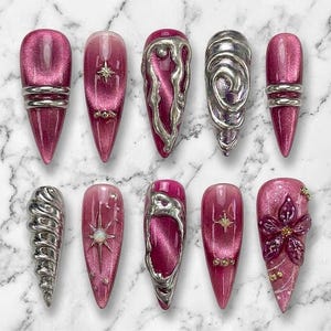 May include: A set of ten press-on nails featuring a deep magenta cat-eye finish. The designs include silver metallic 3D liquid-style accents, raised swirls, star-shaped charms, small embedded rhinestones, and a sculpted magenta floral detail.