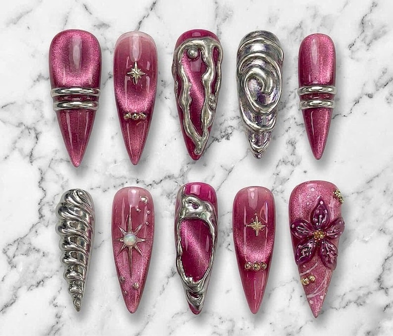 May include: A set of ten press-on nails featuring a deep magenta cat-eye finish. The designs include silver metallic 3D liquid-style accents, raised swirls, star-shaped charms, small embedded rhinestones, and a sculpted magenta floral detail.