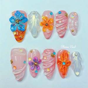 Puede incluir: Diez puntas de uñas rosas y blancas con varios diseños. Algunas puntas presentan flores 3D en azul, naranja y morado. Otras puntas tienen un diseño de espiral con acentos dorados y blancos. El texto "Hana Nail" es visible en la imagen.