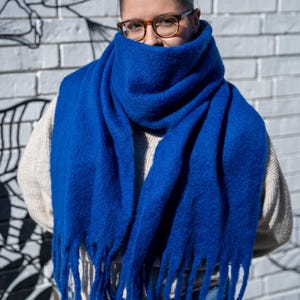 May include: A vibrant blue scarf with fringed edges. The scarf is wrapped around the neck and face, creating a cozy and stylish look. The scarf is made of a soft, textured material. The background is a white brick wall with a black and white mural.