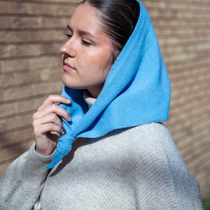 May include: A light blue headscarf tied at the neck. The head covering is made of a soft material and is worn over a light gray sweater. The headscarf is a solid color and is draped over the head.