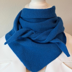 May include: A vibrant blue knit scarf, draped and knotted. The scarf is displayed on a white mannequin head and a white jacket. The scarf's texture is visible, and the color is a rich, solid blue.
