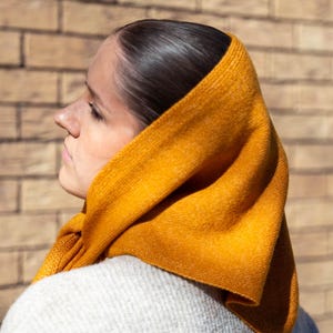 May include: A mustard yellow knitted hood, draped over a light grey sweater. The hood is wrapped around the head and neck, with soft folds. The background is a brick wall.
