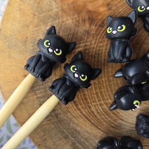 Peut inclure: Des capuchons de crayon en forme de chat noir avec des yeux jaunes et des nez roses. Les capuchons sont fixés à des crayons en bois de couleur claire. D'autres capuchons de chat noir sont éparpillés sur une surface en bois. Accessoire amusant.