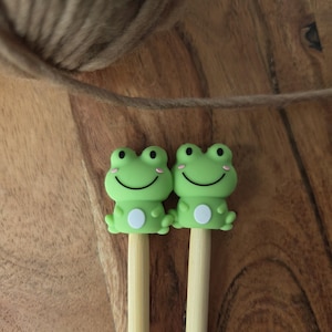 Peut inclure: Deux capuchons de crayon en forme de grenouille verte avec des visages souriants et des ventres blancs. Les capuchons sont fixés à des crayons en bois de couleur claire. Le fond est une surface en bois avec un brin de fil de couleur beige.