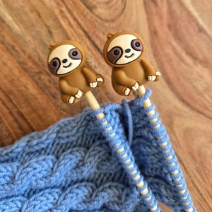 May include: Two knitting needles with brown sloth toppers are inserted into a blue knitted fabric. The sloths have white faces, brown bodies, and black eyes. The knitting project is on a wooden surface.