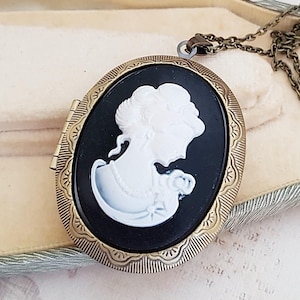 Könnte beinhalten: Eine goldfarbene ovale Halskette mit einem schwarzen Kamee, der eine weiße Silhouette eines Frauenprofils zeigt.