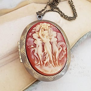 Puede incluir: Un collar con un medallón de camafeo de estilo vintage con un camafeo rojo y blanco con tres mujeres. El medallón es de latón y tiene una cadena.