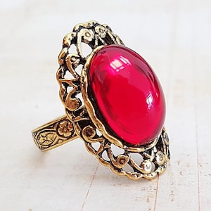Peut inclure: Une bague de style vintage avec une grosse pierre rouge rubis ovale. La bague a un sertissage orné de style filigrane de couleur dorée. La bande présente un motif floral. Un bijou de caractère.
