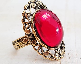 May include: A vintage-style ring with a large, oval, ruby-red gemstone. The ring has an ornate, filigree-style setting in a gold tone. The band features a floral design. A statement piece of jewelry.
