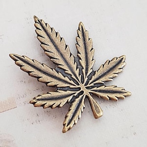 Puede incluir: Un elemento decorativo de metal color bronce en forma de hoja. La hoja tiene vetas detalladas y una superficie texturizada. El objeto es probablemente un broche, colgante u otro componente de joyería. El fondo es claro y texturizado.