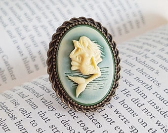 May include: A cameo ring with a bronze setting and a blue and white cameo of a mermaid. The cameo is oval shaped and has a detailed design.
