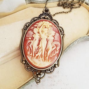 Può includere: Una collana con cammeo con una catena in bronzo e un ciondolo ovale con cammeo con un designo intagliato di tre donne in uno schema di colori bianco e arancione.