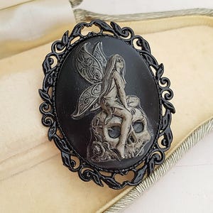May include: Oval black brooch featuring a fairy design seated on a skull, with a black filigree border. The brooch is presented in a cream-coloured jewellery box.