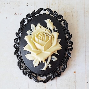 May include: An oval black cameo brooch with a cream rose design. The rose is centrally placed on a black backdrop, framed by an ornate black border. This brooch is a vintage-style accessory, suitable for various outfits.