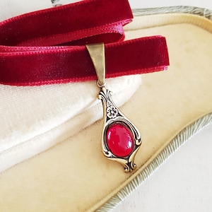 May include: A deep red velvet choker necklace with a decorative pendant. The pendant has an oval, red gemstone set in an ornate, silver-toned metal frame. The necklace is presented in a white jewellery box.
