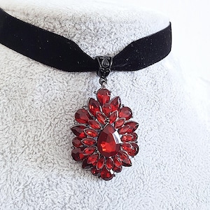 Könnte beinhalten: Ein schwarzes Samt-Choker-Halsband mit einem großen, verzierten Anhänger. Der Anhänger zeigt eine Ansammlung roter Edelsteine in einem floralen Design, mit einem großen, tropfenförmigen Stein in der Mitte. Das Metall ist dunkel.