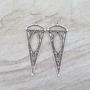 May include: A pair of silver dangle earrings with a geometric, triangular design. The earrings have a detailed, intricate pattern.