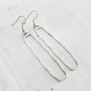 Puede incluir: Un par de pendientes colgantes rectangulares plateados. Los pendientes tienen una textura martillada y están suspendidos de ganchos para las orejas. El diseño es minimalista y moderno, adecuado para el uso diario.