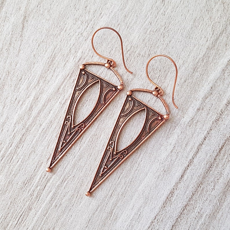 May include: A pair of copper triangle earrings with intricate geometric designs. The earrings have a delicate, openwork pattern and a slightly oxidized finish.
