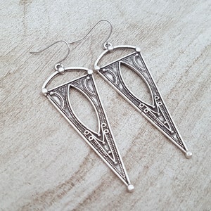 May include: A pair of silver dangle earrings with a geometric triangle design. The earrings have a detailed pattern and a silver hook closure.