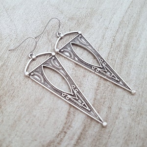 May include: A pair of silver triangle earrings with intricate geometric designs. The earrings have a dangling design and are made of metal.