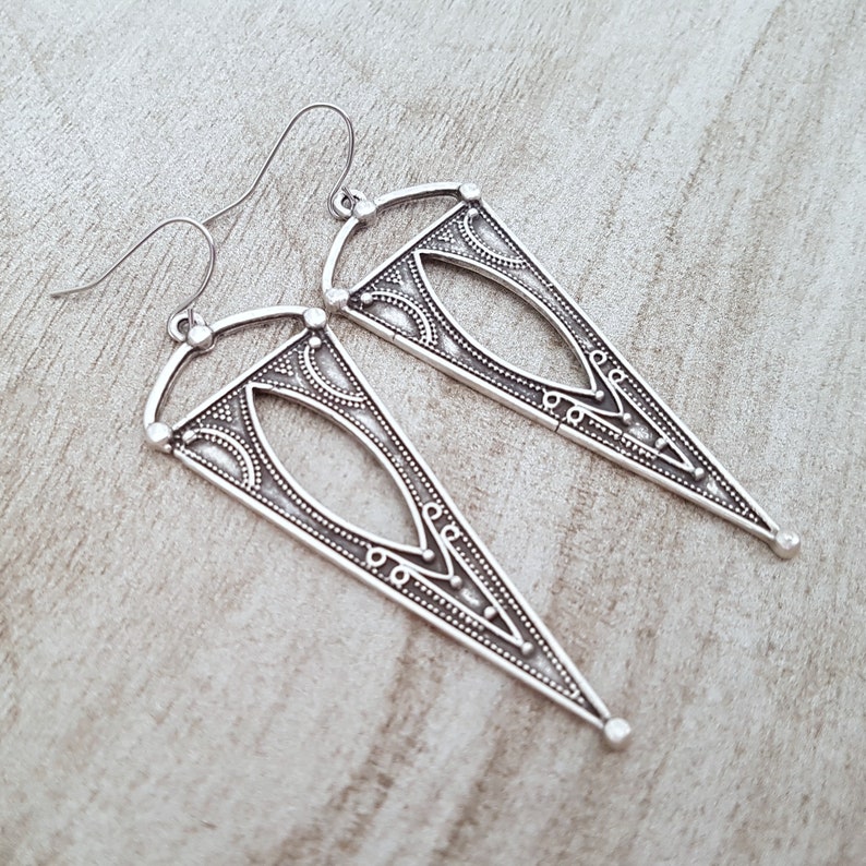 May include: A pair of silver triangle earrings with intricate geometric designs. The earrings have a dangling design and are made of metal.