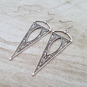 May include: A pair of silver triangle earrings with intricate geometric designs. The earrings have a dangling design and are made of metal.