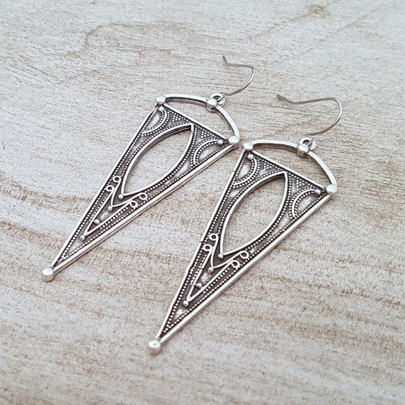 May include: A pair of silver triangle earrings with intricate geometric designs. The earrings have a dangling design and are made of metal.