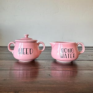 May include: Two pink ceramic pieces, a lidded sugar bowl and a creamer, sit side-by-side. The sugar bowl has the word "WEED" in black lettering, and the creamer reads "BONG WATER". Both pieces have handles and are on a dark wooden surface.