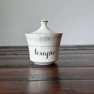 May include: A white porcelain sugar bowl with a lid, featuring a delicate blue floral pattern and silver trim along the rim. The word lexapro is printed in black cursive script on the front of the bowl, which has a tapered base and a knobbed lid.