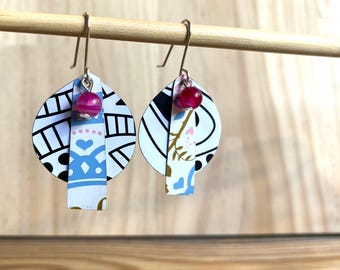 May include: A pair of dangle earrings with a white, black, and blue patterned circle and a pink bead. The earrings are hanging from silver hooks.