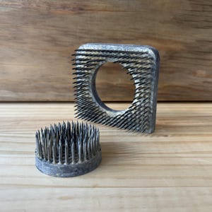 Op de afbeelding: Twee vintage metalen bloemensponzen, één rond en één vierkant met een centraal gat. Beide hebben dicht op elkaar geplaatste, scherpe metalen pinnen voor het schikken van bloemen. De sponzen zijn grijs en liggen op een lichtbruin houten oppervlak.