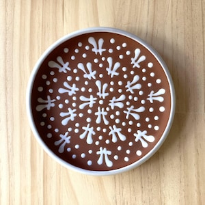 Puede incluir: Un plato de cerámica blanco con un esmalte marrón y un patrón geométrico blanco. El patrón está formado por pequeños puntos y formas de flores estilizadas.