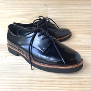 May include: A pair of black leather Oxford shoes with black laces. The shoes have a glossy finish and a brown and black sole. The shoes are displayed on a light wood surface.